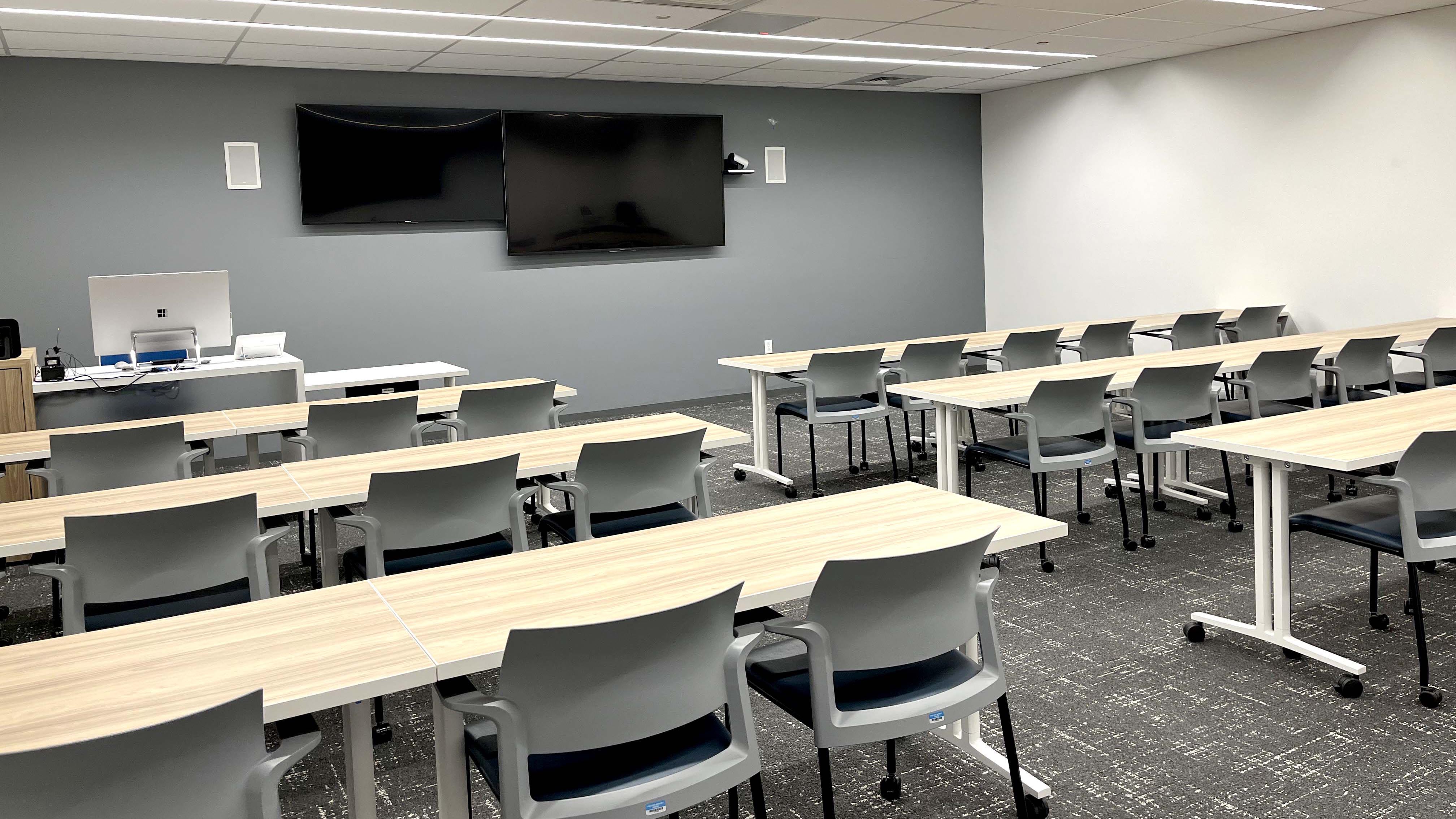 Room with three rows of tables and 30 chairs facing a pair of TVs mounted on the front wall.