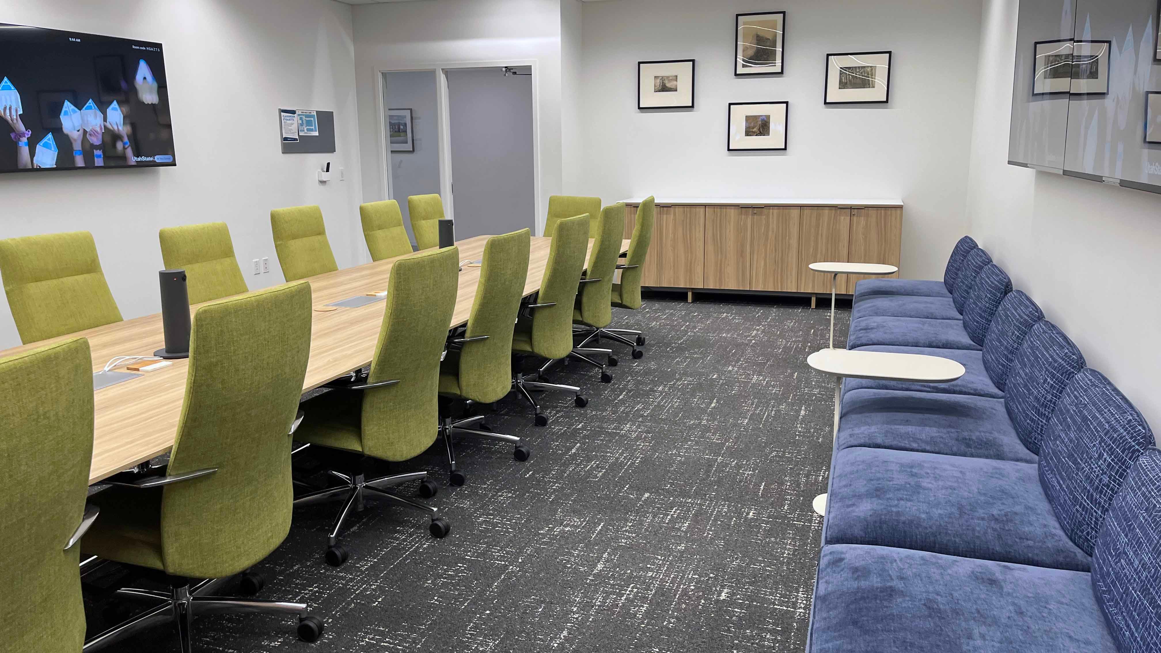 Image of room 159 in the USU Salt Lake Campus showing a large central conference table with 15 chairs, four TVs on surrounding walls, and a long couch on one side of the room.