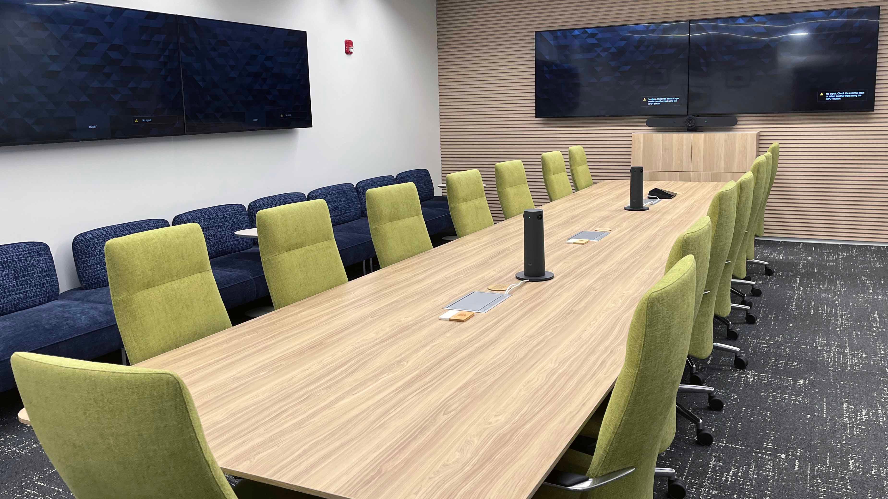 Image of room 159 in the USU Salt Lake Campus showing a large central conference table with 15 chairs, four TVs on surrounding walls, and a long couch on one side of the room.