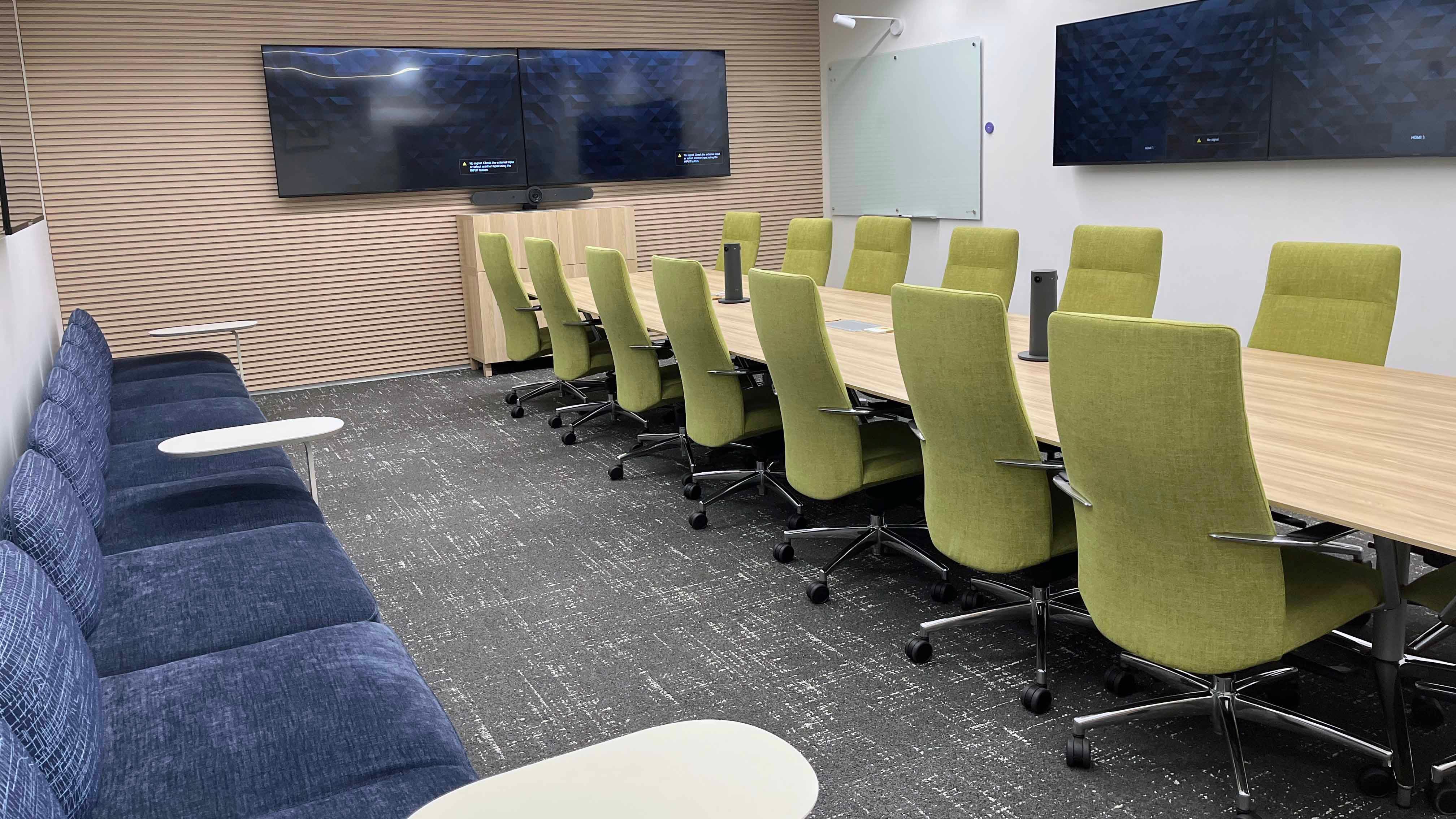 Image of room 159 in the USU Salt Lake Campus showing a large central conference table with 15 chairs, four TVs on surrounding walls, and a long couch on one side of the room.