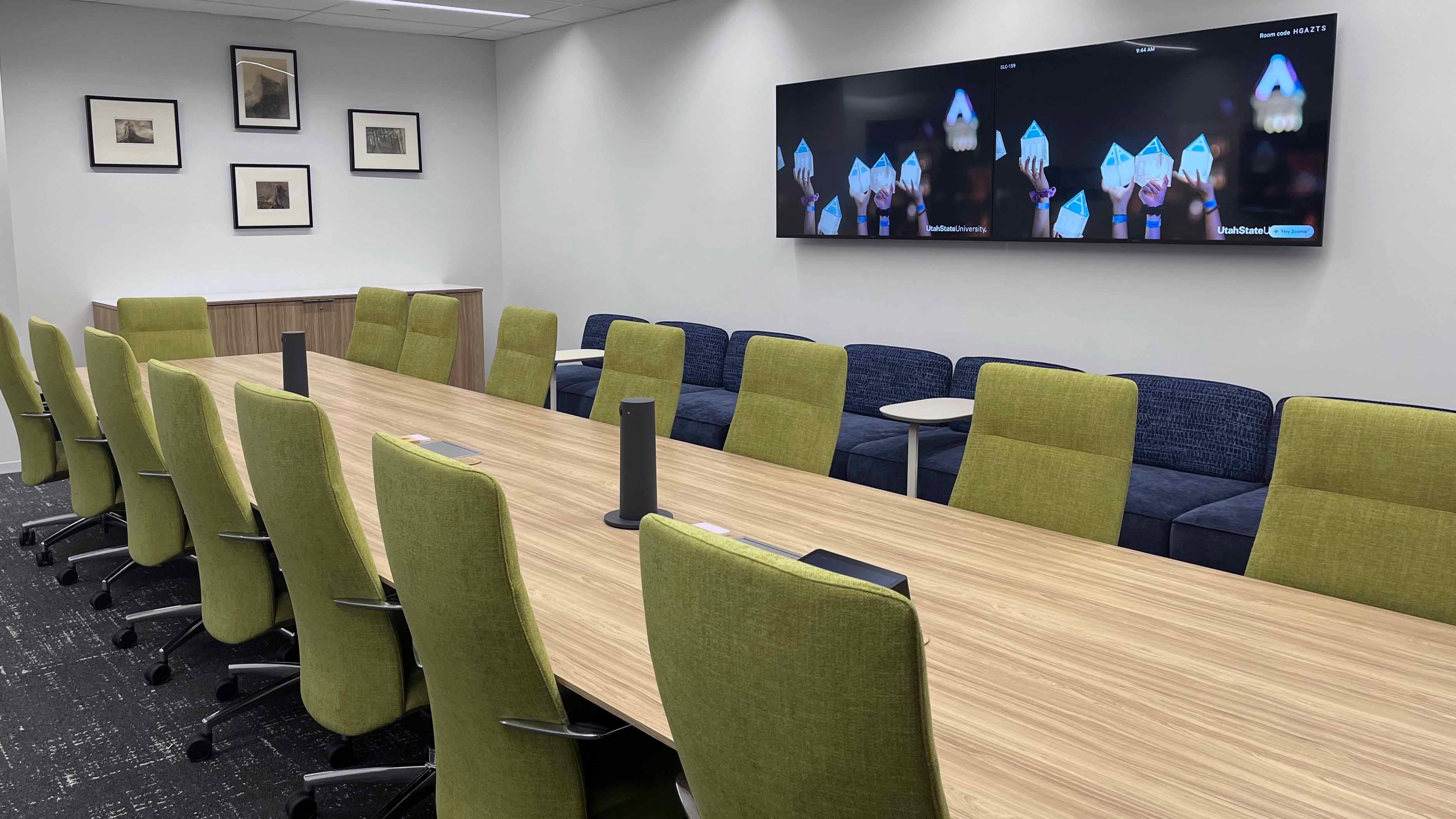 Image of room 159 in the USU Salt Lake Campus showing a large central conference table with 15 chairs, four TVs on surrounding walls, and a long couch on one side of the room.